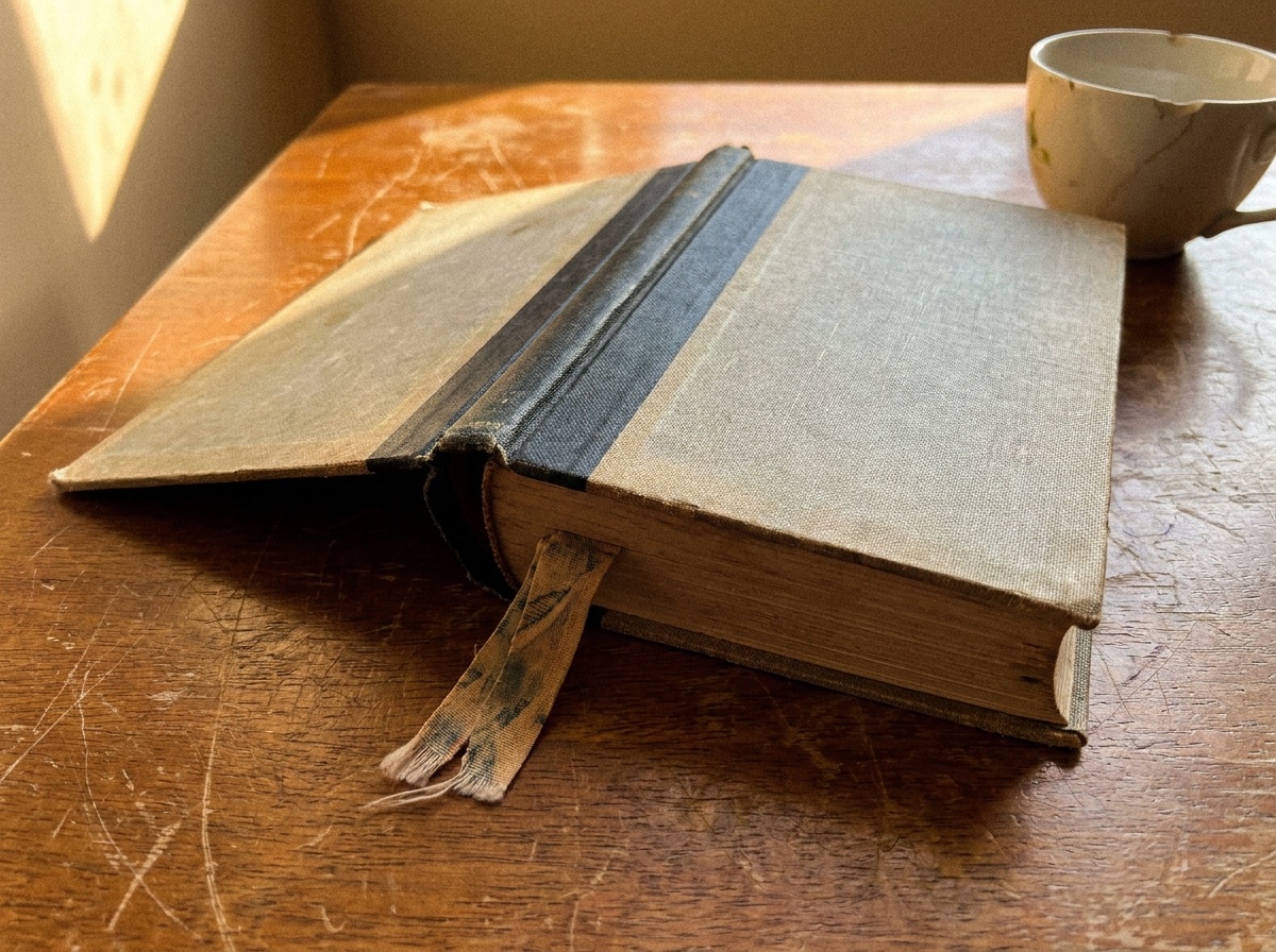 A hardcover book face-down on a wooden table, pages slightly fanned, a small ceramic teacup beside it in soft afternoon window light.